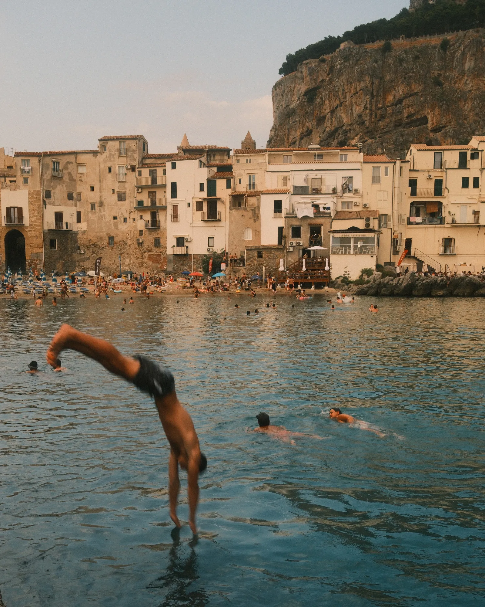 A man jumping into the water in front of the old town of Cefalù at golden hour