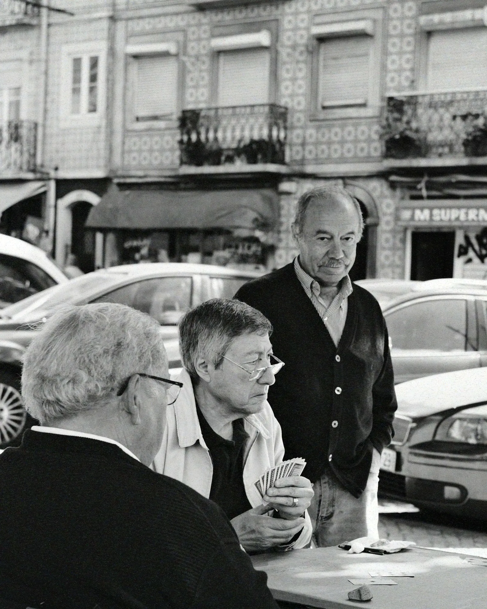Three older men playing cards at a street table in Lisbon, tiled facades and parked cars behind them