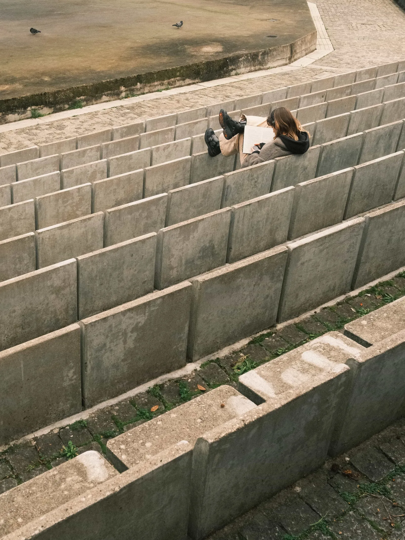 Woman reading a book alone on the concrete steps of an open-air amphitheatre, pigeons on the empty stage