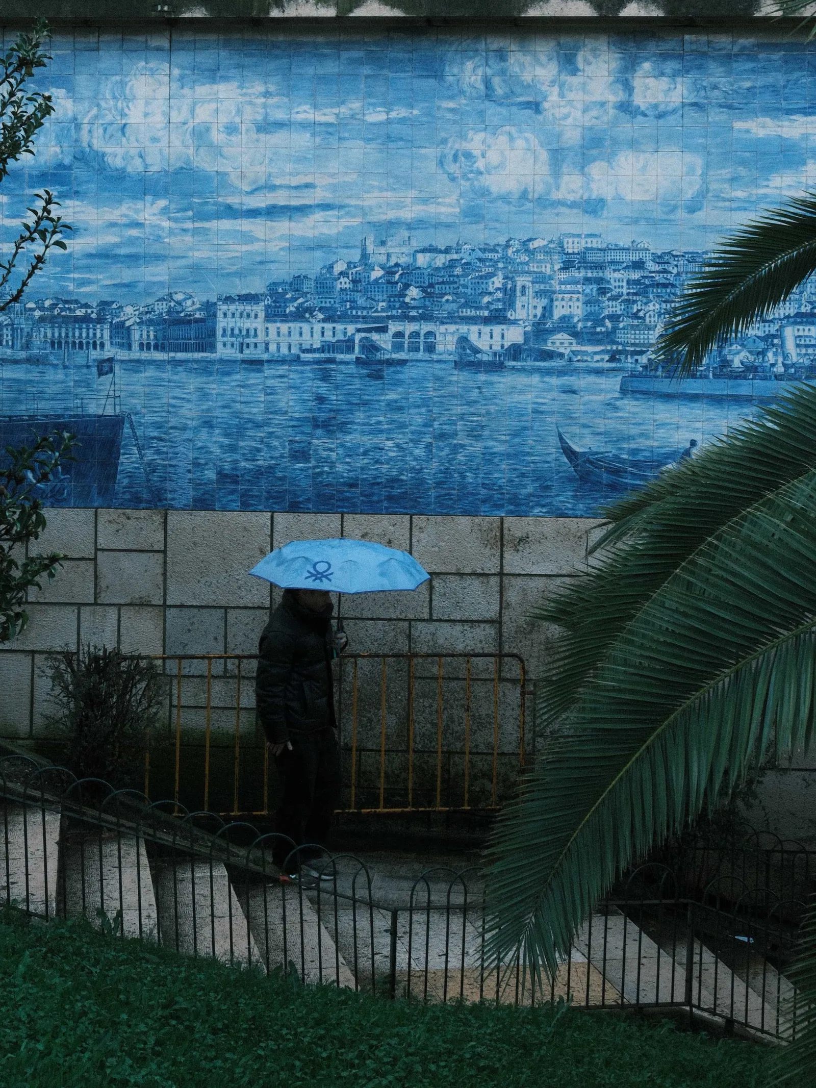 Person under a blue umbrella walking past a large azulejo mural of the Lisbon waterfront, palm frond in the foreground