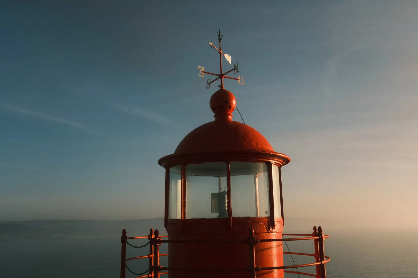 Red lantern room and weather vane of the Nazare lighthouse against a hazy blue and gold sky
